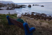 Вид спереду вдумлива активна старша жінка тримає цифровий планшет, сидячи на дерев'яній лавці в морі — стокове фото