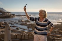 Vue de face d'une femme âgée active prenant un selfie avec son téléphone portable tout en s'appuyant contre un garde-corps sur une promenade le soir pour le coucher du soleil — Photo de stock