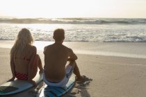Vista trasera de la pareja relajándose en la tabla de surf en la playa en un día soleado. Están viendo las olas. - foto de stock