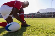 Vista laterale di un giocatore di rugby afroamericano maschile che si lega i lacci delle scarpe sul terreno di rugby sotto il sole — Foto stock