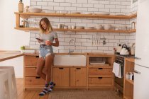Vista frontal de la mujer de pie y el uso de teléfono móvil en casa en la sala de cocina - foto de stock
