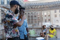 Vista lateral de amigos masculinos multiétnicos brindando con botellas de cerveza en casa en el balcón mientras las mujeres hablan entre sí - foto de stock