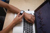 Высокий угол обзора деловой женщины, использующей свои умные часы во время работы со своим ноутбуком за столом в офисе — стоковое фото
