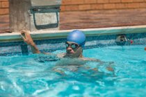 Vista frontal de un joven nadador caucásico que se aferra al borde de la piscina al aire libre mientras usa gorra de natación y gafas en un día soleado - foto de stock