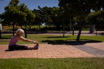 Vista lateral de una mujer mayor activa haciendo ejercicio y estirándose en el parque en un día soleado - foto de stock