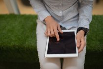 Seção intermediária de uma jovem empresária tocando em um tablet digital enquanto se senta em um banco de grama sintética no escritório — Fotografia de Stock