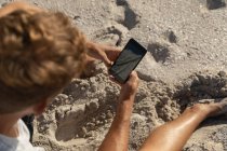 Високий кут зору розслабленого чоловіка, який використовує мобільний телефон на пляжі в сонячний день. Він стоїть — стокове фото