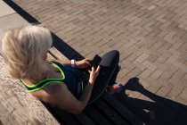 Blick aus der Vogelperspektive auf eine aktive Seniorin, die ihr Handy benutzt und auf einer Bank an der Promenade in der Sonne sitzt — Stockfoto
