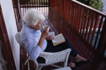 Vista de ángulo alto de una mujer mayor activa tomando café y sentada en el balcón mientras lee un libro en casa - foto de stock