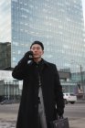 Low angle view of young Asian businessman talking on mobile phone while standing on street with building behind him in the city — Stock Photo