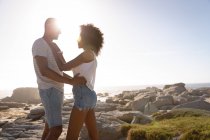 Vista lateral de la pareja afroamericana abrazándose mientras están de pie cerca del lado del mar en la roca - foto de stock