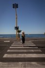 Front view of an active senior woman jogging on the pedestrian crossing of a road next to the sea under the sunshine — Stock Photo
