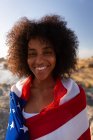 Vista frontal de la mujer afroamericana de pie y envuelta bandera americana cerca del lado del mar mientras mira la cámara - foto de stock