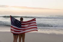 Vista trasera de una joven pareja con bandera americana en la playa. Están mirando el océano. - foto de stock