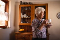 Vista lateral de una mujer mayor activa reflexiva bebiendo su café mientras está de pie en la cocina en casa - foto de stock