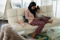 Vista frontal da mãe de raça mista vestindo hijab e filha sentado e usando tablet digital na sala de estar em casa — Fotografia de Stock