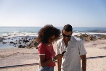 Vista frontal do casal afro-americano usando telefone celular e sorrindo na praia em um dia ensolarado — Fotografia de Stock