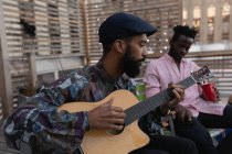 Seitenansicht eines afrikanisch-amerikanischen Mannes, der Gitarre spielt, während ein anderer Freund zu Hause auf dem Balkon ein kaltes Getränk trinkt — Stockfoto