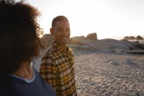 Vista lateral de la pareja afroamericana sonriendo y mirándose cerca del lado del mar. Están parados sobre sable al atardecer. - foto de stock
