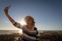 Вид активной пожилой женщины, делающей селфи со своим мобильным телефоном на фоне заката на пляже — стоковое фото