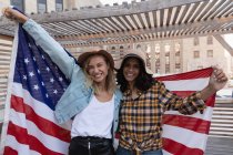 Front view of multi ethnic female friends holding american flag in balcony at home — Stock Photo