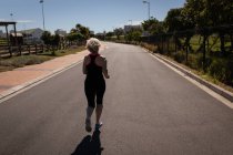 Задний вид активной пожилой женщины, бегущей по пустынной дороге под солнцем — стоковое фото