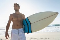 Vista de bajo ángulo del joven surfista masculino con una tabla de surf de pie en la playa en un día soleado. Él está disfrutando de sus vacaciones - foto de stock