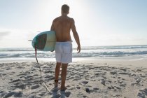Joven surfista masculino con una tabla de surf de pie en una playa en un día soleado. Él está observando las olas - foto de stock
