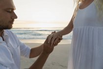 Vista lateral del hombre guapo poniendo anillo en el dedo de mujer en la playa. Él le pregunta en el compromiso - foto de stock
