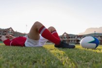 Vue latérale d'un joueur de rugby caucasien contrarié couché avec une balle de rugby dans le stade — Photo de stock