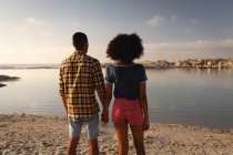 Вид сзади на афро-американскую пару, стоящую на пляже на песке. Они смотрят на море. — стоковое фото
