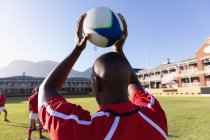 Visão traseira do jogador de rugby americano africano segurando bola sobre sua cabeça para jogar em contato no estádio em um dia ensolarado — Fotografia de Stock