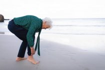 Seitenansicht einer aktiven Seniorin, die die Hosenärmel hochkrempelt, während sie am Strand am Wasser steht — Stockfoto