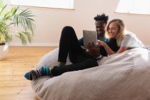 Вид спереду багатоетнічної пари посміхається, сидить і використовує цифровий планшет вдома на дивані — стокове фото
