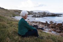 Вид сбоку активной пожилой женщины, сидящей на траве на пляже и смотрящей на море — стоковое фото