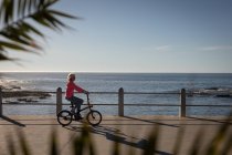 Seitenansicht einer aktiven Seniorin, die am Abend auf einer Strandpromenade Fahrrad fährt — Stockfoto