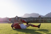 Vue latérale d'un joueur de rugby blessé couché sur le sol et tenant sa cheville au soleil — Photo de stock