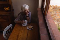 Blick aus der Vogelperspektive auf eine nachdenkliche, aktive Seniorin, die beim Kaffee in der heimischen Küche aus dem Fenster schaut — Stockfoto