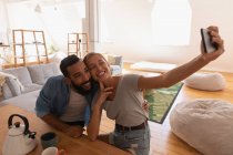 Vue de face du couple mixte prenant selfie dans le salon à la maison — Photo de stock