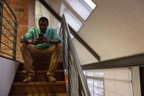 Vue de face de l'homme d'affaires afro-américain utilisant un téléphone portable à l'escalier dans le bureau — Photo de stock