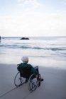 Visão traseira de uma mulher idosa ativa deficiente olhando para o mar enquanto sentada em uma cadeira de rodas na praia — Fotografia de Stock