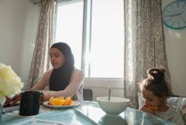 Vista de ángulo bajo de la madre de raza mixta que usa hijab usando computadora portátil mientras su hija come fruta en casa - foto de stock