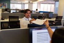 Vista frontal de guapos ejecutivos masculinos dando trozo de papel a una colega caucásica en un escritorio en una oficina moderna - foto de stock