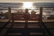 Вид сзади активной пожилой женщины, расслабляющейся на скамейке и смотрящей на закат перед пляжем — стоковое фото