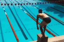 Rear view of young Caucasian male swimmer standing on starting block in starting position at outdoor swimming pool on sunny day — Stock Photo