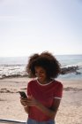 Vista frontale della bella donna afro-americana sorridente mentre si utilizza il telefono cellulare in spiaggia — Foto stock