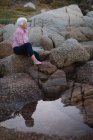 Seitenansicht einer nachdenklichen aktiven kaukasischen Seniorin, die am Strand auf einem Felsen sitzt — Stockfoto