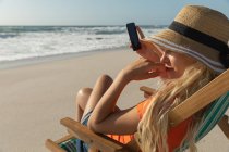 Vista lateral da mulher relaxando na espreguiçadeira na praia em um dia ensolarado. Ela está sentada e usando seu telefone celular — Fotografia de Stock