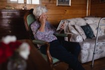Seitenansicht einer aktiven Seniorin mit der Hand an der Schläfe, die ein digitales Tablet benutzt, während sie zu Hause auf einem Sessel sitzt — Stockfoto