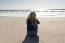 Вид сзади на красивую блондинку, сидящую на пляже в солнечный день. Она смотрит в пейзаж — стоковое фото